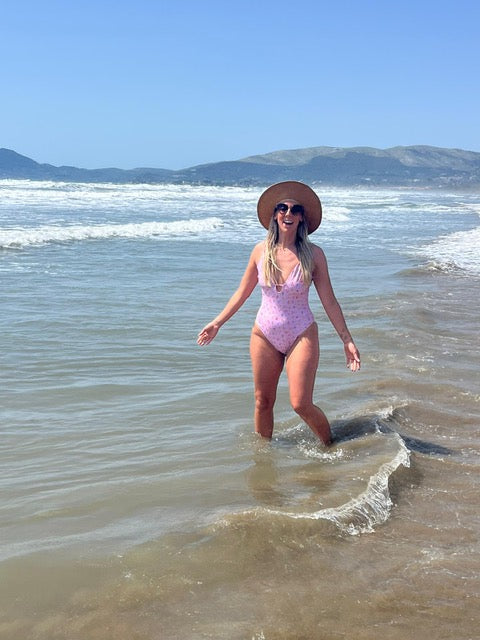 Woman in a pink swimsuit and straw hat standing in shallow water at the beach with mountains in the background.