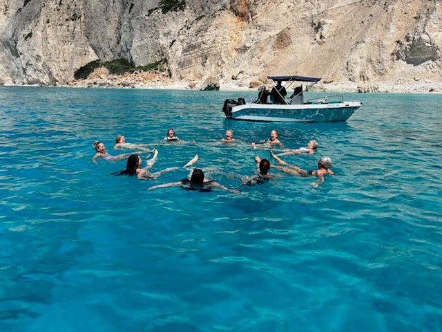 People floating on their backs forming a circle in the clear blue sea with a boat and white cliffs in the background