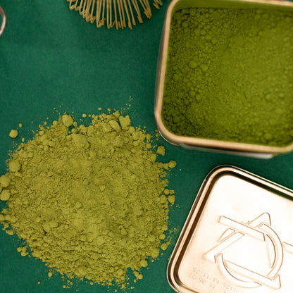 Vibrant green ceremonial matcha powder spilled next to an open 50g tin and a bamboo whisk on a green surface.