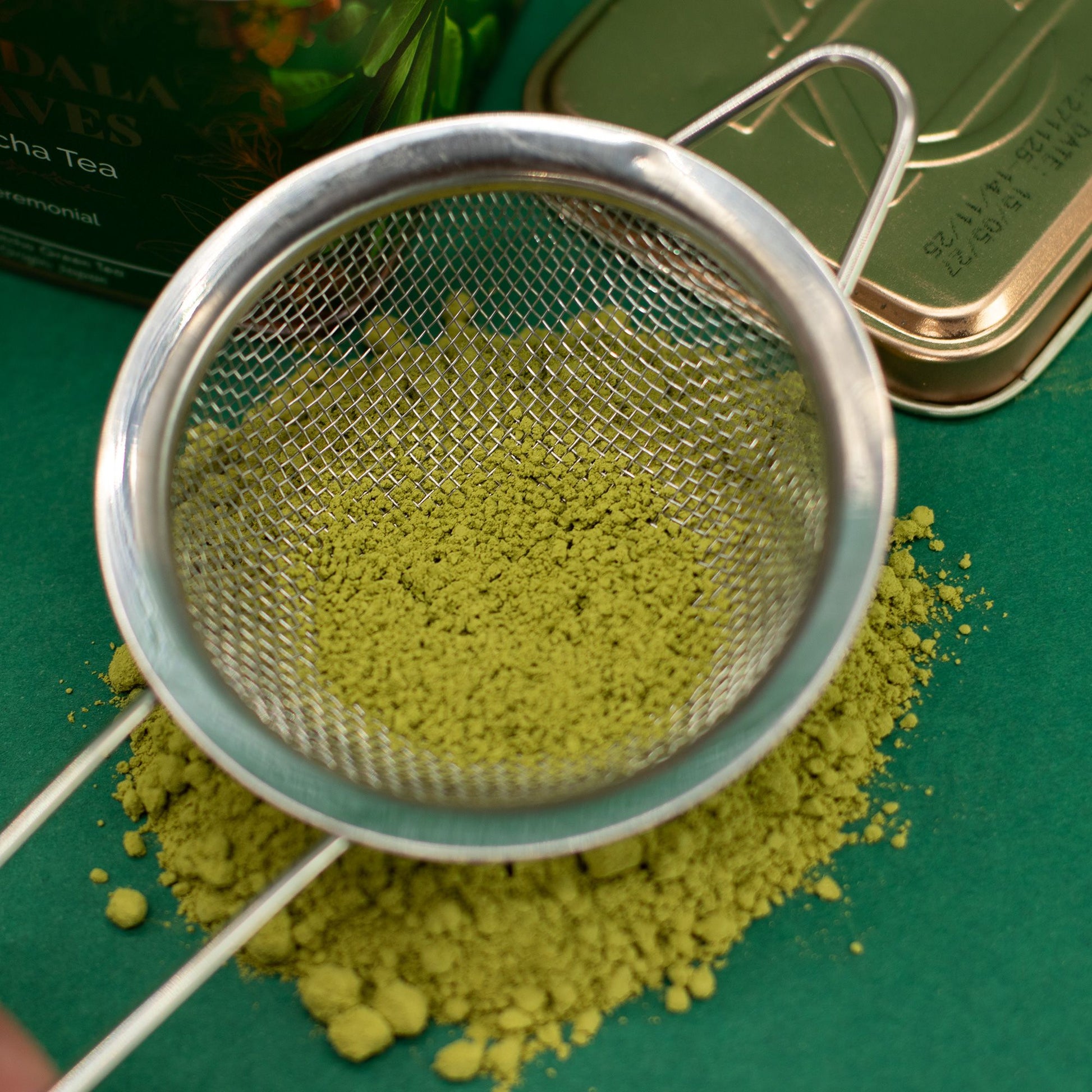 Vibrant green ceremonial matcha tea powder in a silver mesh sifter, with a decorative green tin in the background.