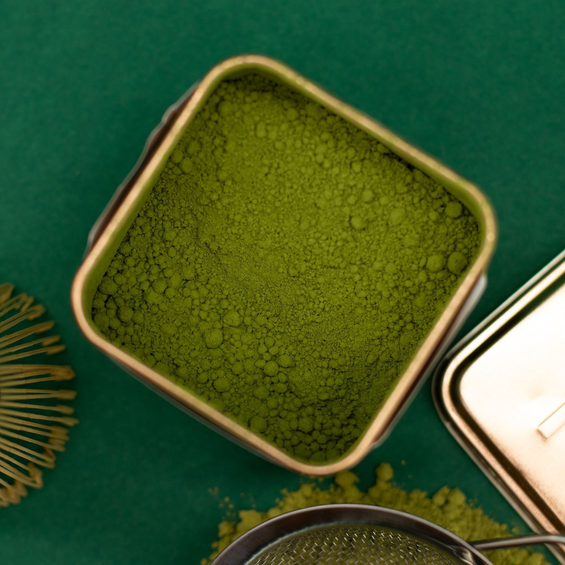 Vibrant green ceremonial matcha powder in an elegant 50g tin, with a whisk and sifter on a green background.