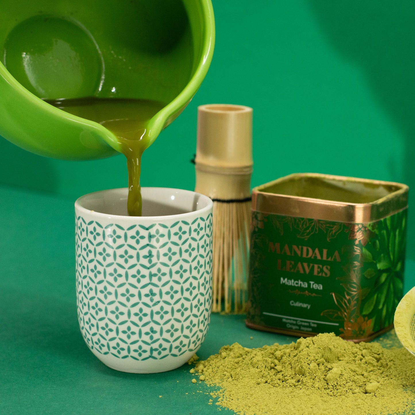 Vibrant green matcha tea being poured into a patterned white cup, with a green Mandala Leaves tin and bamboo whisk.