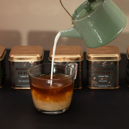Green teapot pouring milk into a glass of iced tea, with black and gold tea tins in the background.