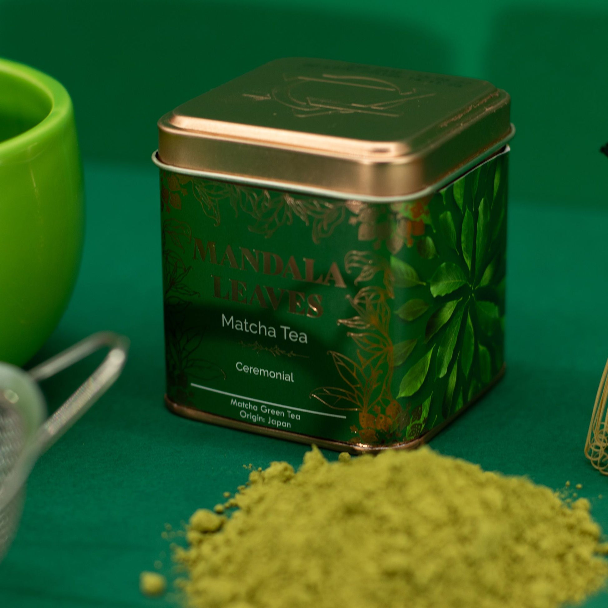 Green Mandala Leaves 50g Ceremonial Matcha Tea tin with matcha powder, whisk, and cup on a green surface.