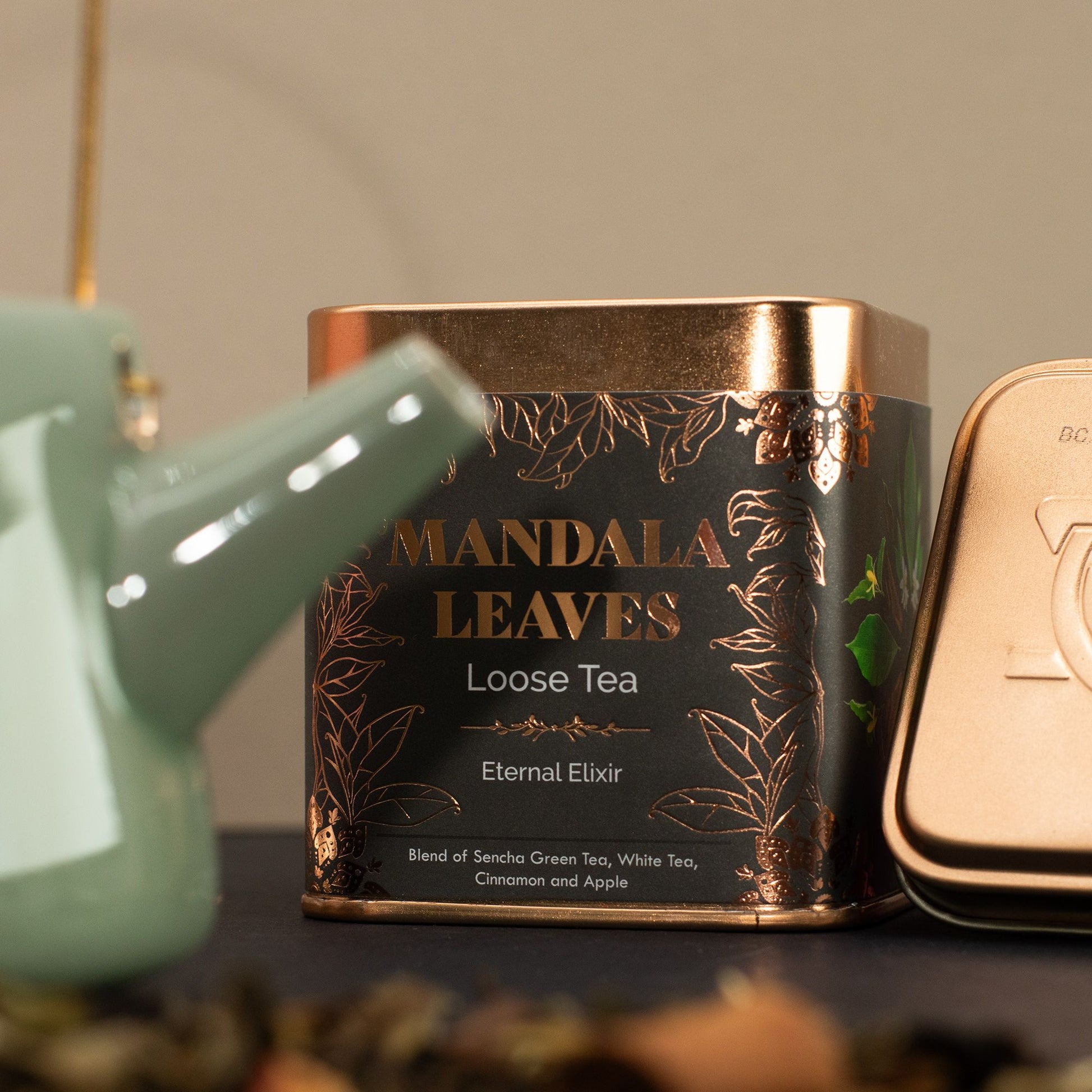 Mandala Leaves Eternal Elixir 50g tea tin with gold and grey label, next to a green teapot.