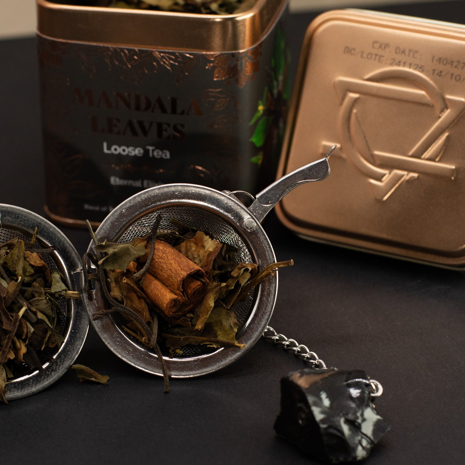 Loose green and white tea with cinnamon sticks in a metal infuser, with two bronze tea tins.