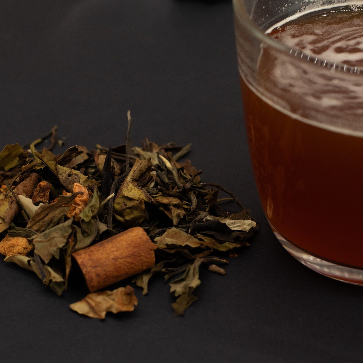 Loose green and white tea leaves with cinnamon stick next to a glass of brewed Eternal Elixir tea.